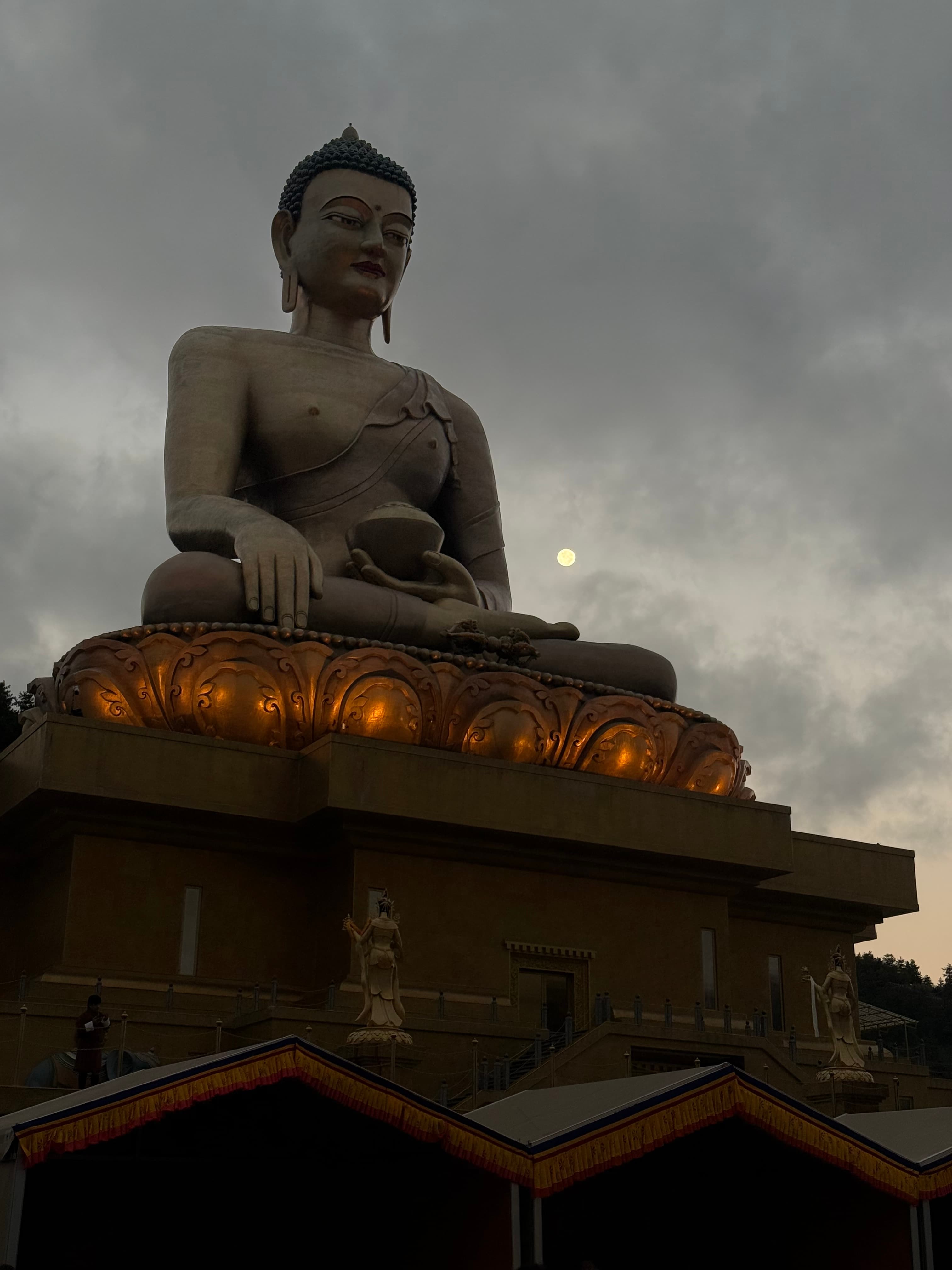 A large Buddha statue silhouetted against clouds, the moon rising just behind it.